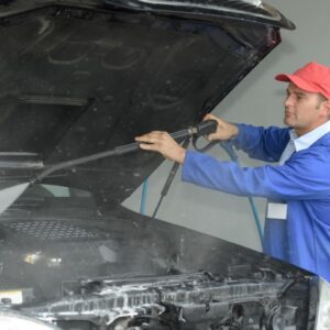 A man in blue jacket holding hose over car engine.