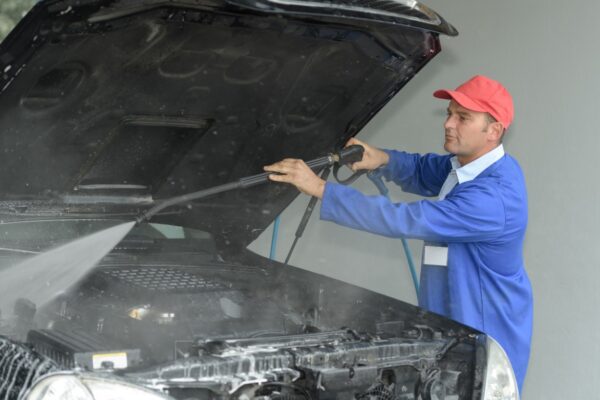 A man in blue jacket holding hose over car engine.